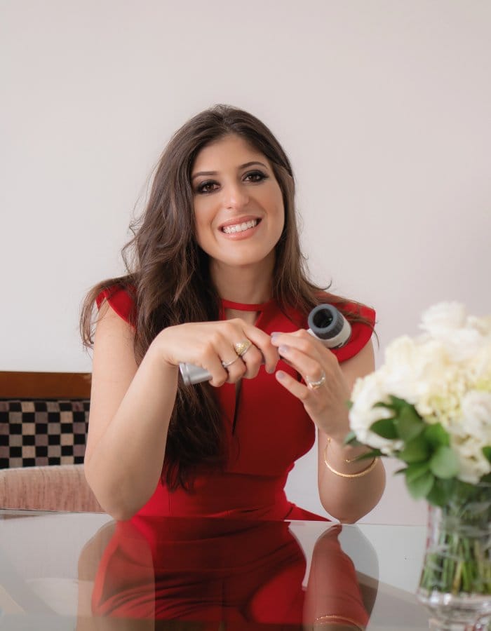 A imagem mostra a Dra. Barbara Tanus sentada à mesa, vestindo um elegante vestido vermelho. Ela está segurando um pequeno objeto cilíndrico em suas mãos, possivelmente um produto dermatológico ou cosmético. Seus longos cabelos castanhos caem sobre os ombros, e ela usa vários anéis e pulseiras, complementando seu visual sofisticado. A mesa é de vidro, e ao lado dela há um vaso com flores brancas, adicionando um toque de elegância ao ambiente. O fundo é simples e claro, destacando a Dra. Barbara Tanus como o foco principal da imagem.