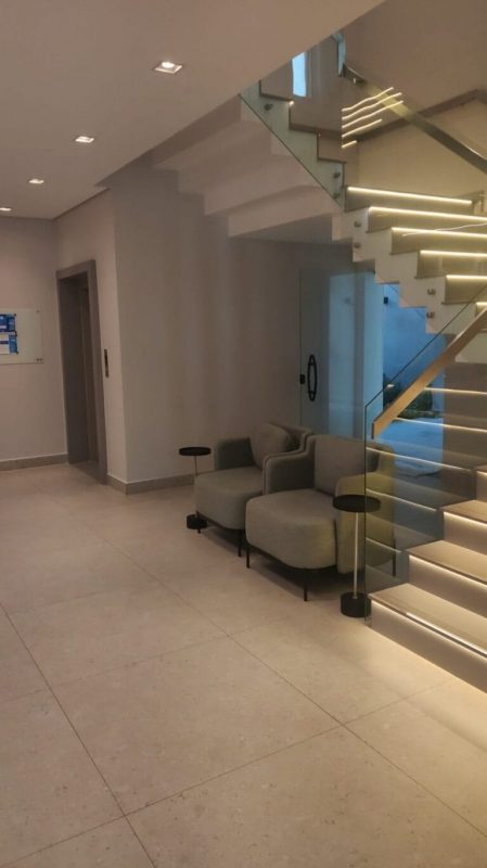 A imagem mostra um moderno hall de entrada com uma escada de design contemporâneo. A escada possui degraus iluminados por fitas de LED, criando um efeito visual atraente e sofisticado. Ao lado da escada, há um pequeno lounge com duas poltronas cinza e duas mesinhas laterais pretas, proporcionando um espaço confortável para espera. O ambiente é minimalista, com paredes brancas e piso de mármore claro, reforçando a sensação de elegância e modernidade. A iluminação embutida no teto contribui para a atmosfera acolhedora e bem iluminada do espaço.